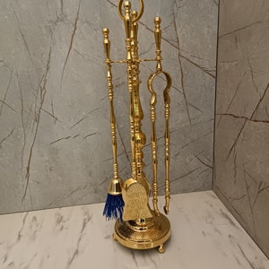 May include: A gold metal fireplace tool set with a brush, shovel, poker, and tongs. The tools are mounted on a decorative stand with a round base.