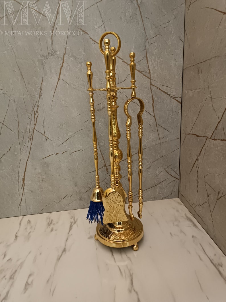 May include: A gold metal fireplace tool set with a brush, shovel, poker, and tongs. The tools are mounted on a decorative stand with a round base.