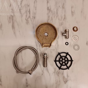 K&ouml;nnte beinhalten: Ein Satz Messing-Armaturen f&uuml;r einen Wasserbrunnen, bestehend aus einer Messing-Schale, einem schwarzen Metallgitter, einem verchromten Schlauch und einem verchromten Ventil. Die Teile liegen auf einer wei&szlig;en Marmorfl&auml;che.