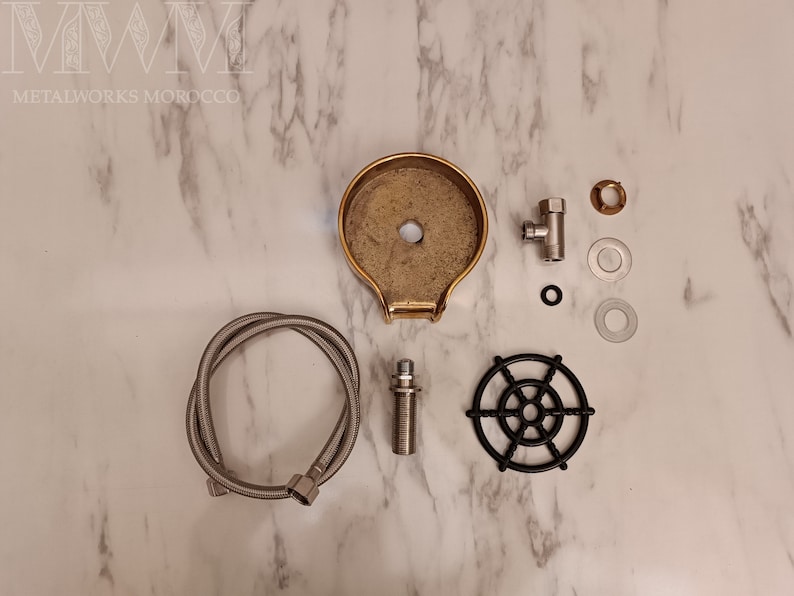 K&ouml;nnte beinhalten: Ein Satz Messing-Armaturen f&uuml;r einen Wasserbrunnen, bestehend aus einer Messing-Schale, einem schwarzen Metallgitter, einem verchromten Schlauch und einem verchromten Ventil. Die Teile liegen auf einer wei&szlig;en Marmorfl&auml;che.