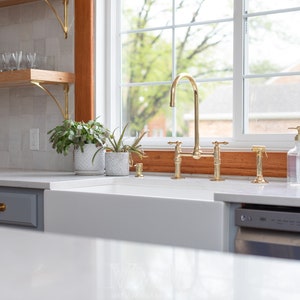 May include: A white farmhouse kitchen sink with a gold faucet and a gray dishwasher. The sink is under a window with a wooden frame. There are plants on the counter next to the sink.