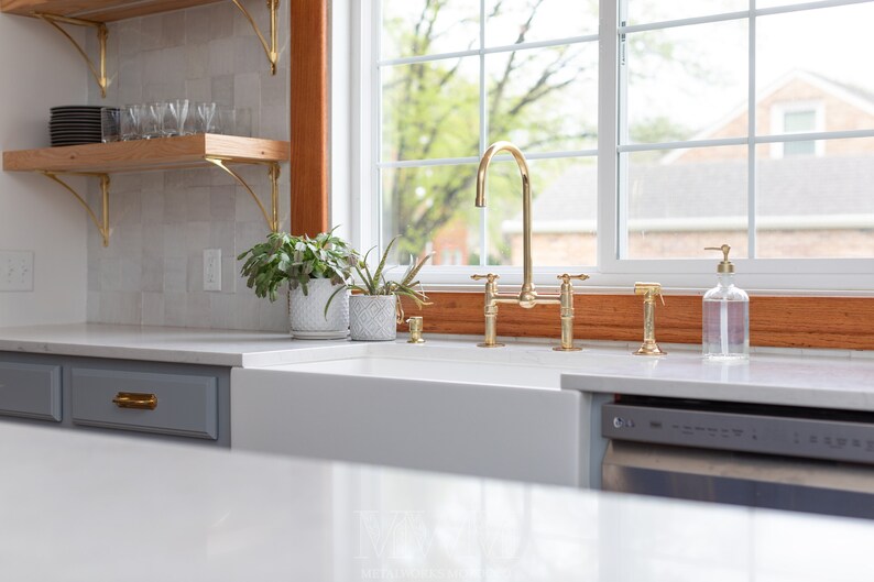 May include: A white farmhouse kitchen sink with a gold faucet and a gray dishwasher. The sink is under a window with a wooden frame. There are plants on the counter next to the sink.