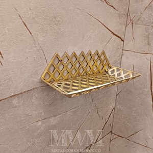 May include: A gold metal shelf with a geometric design. The shelf is mounted on a wall with a light beige background.