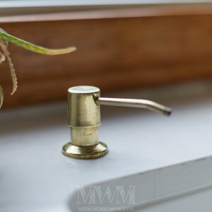 May include: Gold soap dispenser with a pump and a spout. The dispenser is on a white countertop.