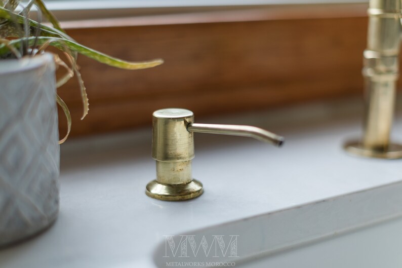 May include: Gold soap dispenser with a pump and a spout. The dispenser is on a white countertop.