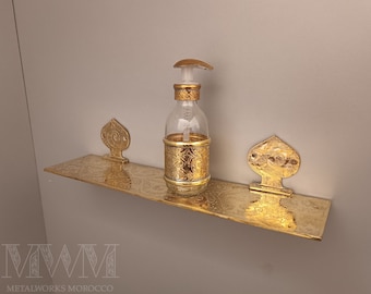 May include: A gold metal shelf with intricate designs, featuring a gold soap dispenser with a clear glass bottle. The shelf is mounted on a wall.