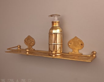 Puede incluir: Un estante de metal dorado con dos soportes decorativos y un dispensador de jabón dorado. El estante está montado en una pared.