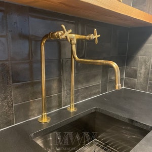 May include: A gold-toned double-handle faucet with a long, curved spout, mounted on a black countertop. The faucet is installed against a backdrop of dark gray tiles.