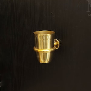 May include: A gold-colored metal toothbrush holder mounted on a black wall. The holder has a round base and a cylindrical cup.