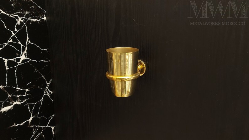 May include: A gold-colored metal toothbrush holder mounted on a black wall. The holder has a round base and a cylindrical cup.