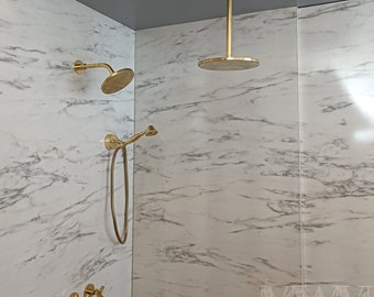 May include: A gold shower head with a round, flat design is mounted on the ceiling. A smaller gold shower head is mounted on the wall, and a handheld shower head is attached to a hose. There are also four gold knobs on the wall. The shower is in a white marble bathroom.