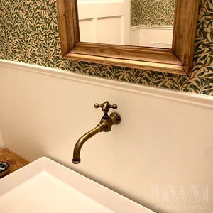 Puede incluir: Un grifo de latón de estilo vintage con un mango en forma de cruz está montado en una pared blanca. El grifo tiene un caño curvo y está situado encima de un lavabo rectangular blanco. Un espejo con marco de madera y papel pintado se ven en el fondo.