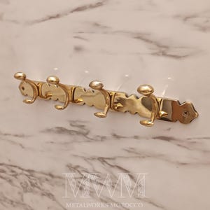 Peut inclure: Un porte-manteau mural en laiton avec quatre crochets. Le porte-manteau a un bord décoratif festonné et une finition polie. Les crochets sont incurvés et ont un bouton arrondi en haut. Le porte-manteau est monté sur un mur à motifs de marbre.