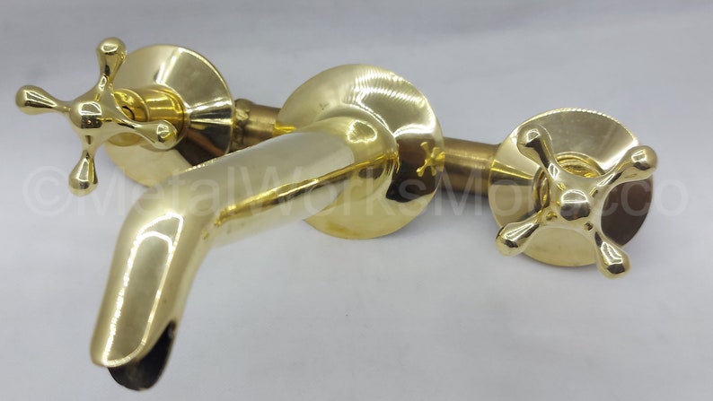 May include: A gold-toned, wall-mounted faucet with two cross-shaped handles. The faucet has a spout that curves downwards.