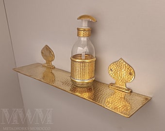 May include: Gold metal shelf with a hammered finish and two decorative spade-shaped brackets. A clear glass soap dispenser with a gold top and a gold textured body sits on the shelf.