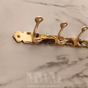 May include: A brass wall-mounted coat rack with four curved hooks. The rack has a decorative, scalloped edge and is mounted on a marble-patterned surface. The text "MWM METALWORKS MOROCCO" is at the bottom.