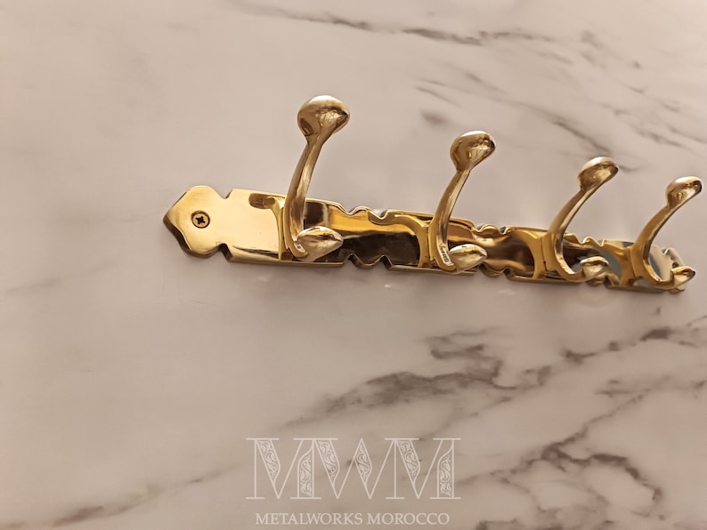 May include: A brass wall-mounted coat rack with four curved hooks. The rack has a decorative, scalloped edge and is mounted on a marble-patterned surface. The text "MWM METALWORKS MOROCCO" is at the bottom.