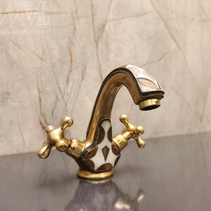 May include: A brass bathroom faucet featuring a curved spout and two cross-shaped handles. The faucet body is decorated with inlaid white and dark brown geometric patterns, and the metal surface has a warm, aged gold-toned finish throughout.