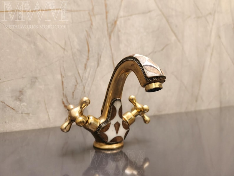 May include: A brass bathroom faucet featuring a curved spout and two cross-shaped handles. The faucet body is decorated with inlaid white and dark brown geometric patterns, and the metal surface has a warm, aged gold-toned finish throughout.