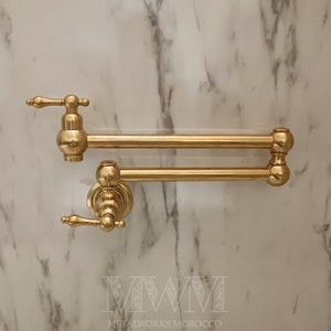 Könnte beinhalten: Ein goldfarbener Doppelarm-Topf-Füller mit einem Hebelgriff. Der Wasserhahn ist an einer weißen Marmorwand montiert. Der Text "MWM METALWORKS MOROCCO" ist am unteren Rand des Bildes sichtbar.