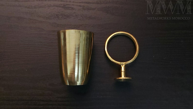 May include: A gold metal cup and a gold metal ring stand on a dark wood surface. The cup has a textured surface. The ring stand has a round top and a base.