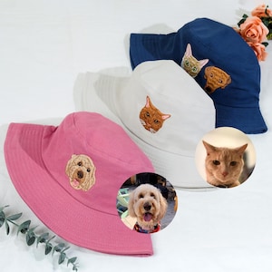 May include: Three bucket hats with embroidered pet portraits. The hats are pink, white, and blue. The portraits are of a dog, a cat, and a cat.