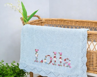 May include: A light blue baby blanket with the name "Lolla" embroidered in floral letters. The blanket has a decorative, scalloped edge and is draped over a wicker bassinet. A small plant and flowers are in the background.