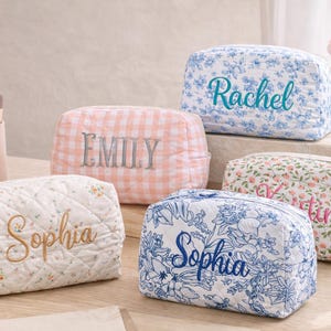 Op de afbeelding: Vijf gepersonaliseerde make-up tassen met bloemen- en ruitpatronen. Elke tas heeft een geborduurde naam: Sophia, Rachel, Kristin en Emily. Make-up borstels en een vaas met tulpen zijn ook zichtbaar.