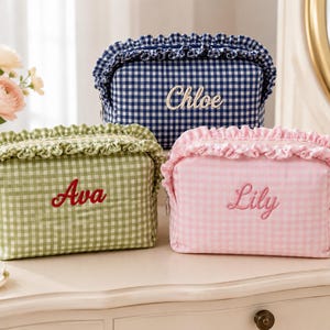 May include: Three rectangular fabric cosmetic bags with ruffled top edges and gingham patterns in green, pink, and navy blue. Each bag features embroidered names on the front: Ava in red, Lily in pink, and Chloe in white script lettering.