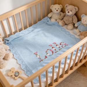 Op de afbeelding: Een lichtblauwe babydeken met de naam "Lola" geborduurd in bloemenletters. De deken heeft een geschulpte rand en ligt in een houten wieg met teddyberen.