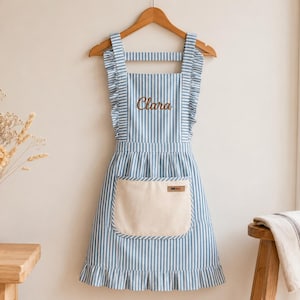 Puede incluir: Un delantal de rayas verticales azules y blancas con volantes en los tirantes y en el dobladillo inferior. El delantal incluye un bolsillo delantero de color crema con una pequeña etiqueta rectangular y el nombre Clara bordado en marrón.