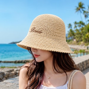 May include: A beige woven straw bucket hat featuring a rounded crown and a downward-sloping brim. The front of the hat is embroidered with the word "Brevete" in dark brown cursive script, showcasing a textured, open-knit construction.