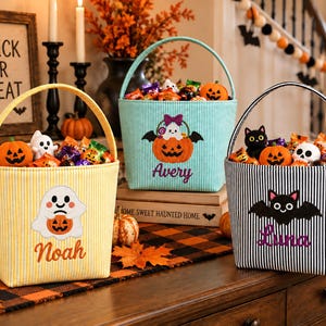 May include: Three fabric Halloween candy buckets with handles, featuring vertical stripes in yellow, light blue, and black. Each bucket is embroidered with a unique character, including a ghost, a pumpkin, and a bat, along with the names Noah, Avery, and Luna.