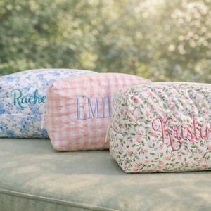 Peut inclure: Trois trousses de maquillage matelassées personnalisées avec des noms brodés. L'une est à fleurs bleues, l'autre à carreaux roses et la troisième à motifs floraux avec des roses roses. Les sacs sont rectangulaires.