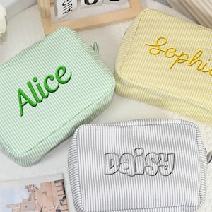 May include: Three rectangular cosmetic bags with striped patterns. One is light green with "Alice" in green script, another is yellow with "Sophia" in yellow script, and the third is gray with "Daisy" in silver script. Each bag has a small loop.