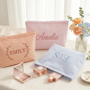 May include: Three rectangular fabric pouches in peach, light pink, and light blue with white pinstripes. The peach pouch features the name EMILY embroidered in a laurel wreath, the pink pouch says Amelia, and the blue pouch features the letters EIS.