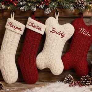 May include: Four hanging Christmas stockings featuring cable knit patterns. Two are traditional stocking shapes in cream and red with names Harper and Chester embroidered. Two are bone-shaped in cream and red with names Buddy and Milo embroidered.