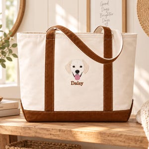 May include: A cream-colored canvas tote bag with brown trim and handles. The front features an embroidered illustration of a golden retriever dog face with its tongue out, positioned above the embroidered brown text that reads Daisy.