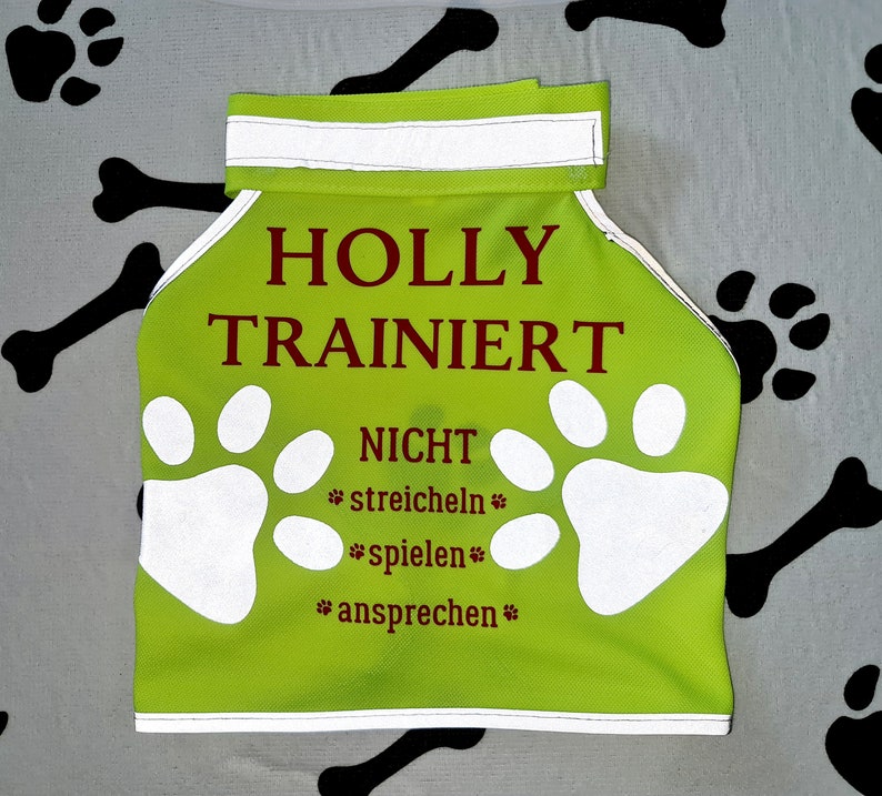 K&ouml;nnte beinhalten: Eine limegr&uuml;ne Hunde-Weste mit reflektierenden wei&szlig;en Pfotenabdr&uuml;cken und dem Text "HOLLY TRAINIERT NICHT *streicheln* *spielen* *ansprechen*" in Braun.