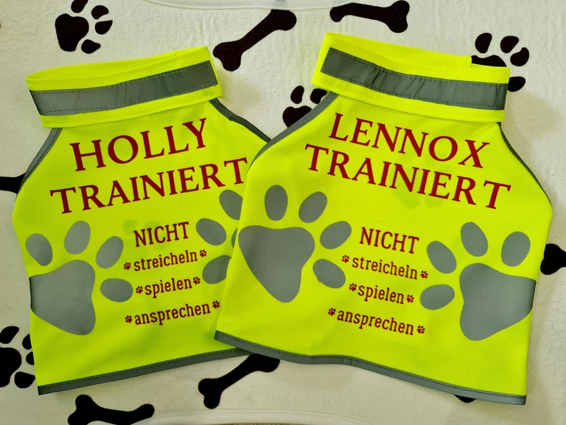 K&ouml;nnte beinhalten: Zwei gelbe Sicherheitswesten mit reflektierenden Streifen. Die Westen haben die Worte "HOLLY TRAINIERT" und "LENNOX TRAINIERT" in Rot aufgedruckt. Die Westen haben auch die Worte "NICHT" und "*streicheln*" in Rot aufgedruckt. Die Westen haben ein Pfotenabdruck-Design.