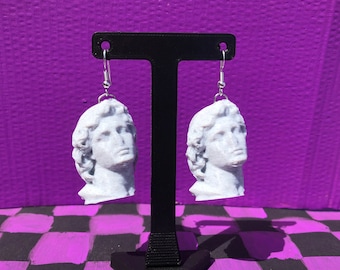 May include: A pair of white, sculpted head earrings with silver hooks. The earrings are displayed on a black stand against a purple background. The heads have detailed facial features and curly hair, giving them a classical appearance.