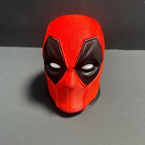 Könnte beinhalten: Eine rot-schwarze Deadpool-Maske mit weißen Augenformen und einer strukturierten Oberfläche. Die Maske ist eine Nachbildung des Gesichts der Comicfigur mit einer schwarzen Umrandung um Augen und Mund.