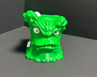 May include: A bright green, 3D-printed planter shaped like a cartoon monster with an angry expression. The planter has a wide mouth, furrowed brow, and white eyes. The design features a textured, layered appearance.