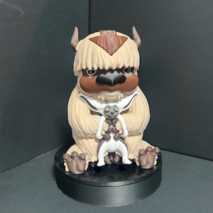 Op de afbeelding: Een gedetailleerd beeldje van Appa, een fictieve vliegende bizon uit Avatar: The Last Airbender. De figuur is voornamelijk beige met bruine accenten, waaronder hoorns en een kenmerkende pijlvormige markering. Een kleiner figuur van Momo staat voor Appa.