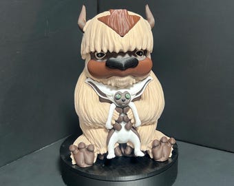 May include: A detailed figurine of Appa, a fictional flying bison from Avatar: The Last Airbender. The figure is primarily beige with brown accents, including horns and a distinctive arrow-shaped marking. A smaller figure of Momo is in front of Appa.