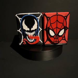Puede incluir: Dos cajas cuadradas pintadas a mano. Una presenta la cara de Venom en negro, azul y rojo, la otra la máscara de Spider-Man en rojo y blanco. Ambas están sobre un soporte negro sobre un fondo negro. Las cajas son probablemente para almacenamiento o exhibición.