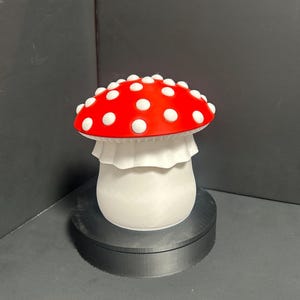 Pode incluir: Estatueta decorativa de cogumelo com chapéu vermelho e pintas brancas. O cogumelo tem um caule branco e uma gola branca com babados. A estatueta é exibida em um suporte circular preto sobre um fundo escuro.