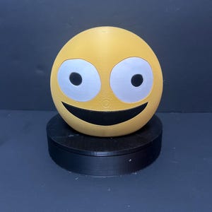 Op de afbeelding: Een gele, bolvormige emoji met grote, zilveromrande ogen en een brede, zwarte glimlach. De emoji staat op een zwarte, cirkelvormige basis tegen een donkere achtergrond. Het ontwerp is eenvoudig en gedurfd, met een cartoonachtige uitstraling.