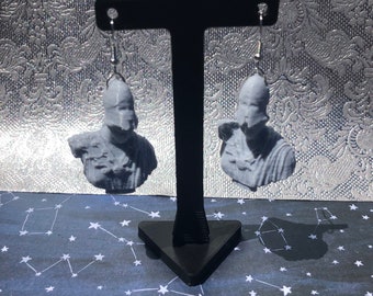 May include: Gray earrings featuring miniature busts of a classical figure. The earrings are suspended from silver-colored hooks. The busts are detailed with facial features and clothing. The earrings are displayed on a black stand against a patterned background.