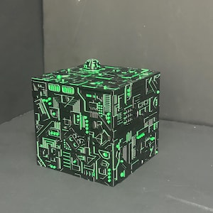 Peut inclure: Un cube noir orné de motifs de circuits imprimés verts en relief sur toutes ses faces. Le design présente des lignes géométriques, des petits carrés et des formes rectangulaires, avec un petit dôme arrondi centré sur la face supérieure.